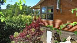 Una cabaña de madera con un ventanal grande, rodeada de abundante vegetación y un árbol de hojas rojas.