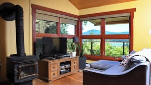 Una sala de estar acogedora con una estufa de leña, un televisor y un sofá con vistas a la vegetación y montañas a través de amplios ventanales.