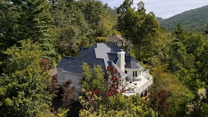 Une maison entourée d’une forêt dense, avec une cheminée imposante et un balcon donnant sur les arbres.
