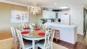 A modern kitchen with white cabinetry, a central island, and a dining area with a glass table and white chairs. - Dolphin Watch 7 Tybee Vacation Rentals (Tybee Island)