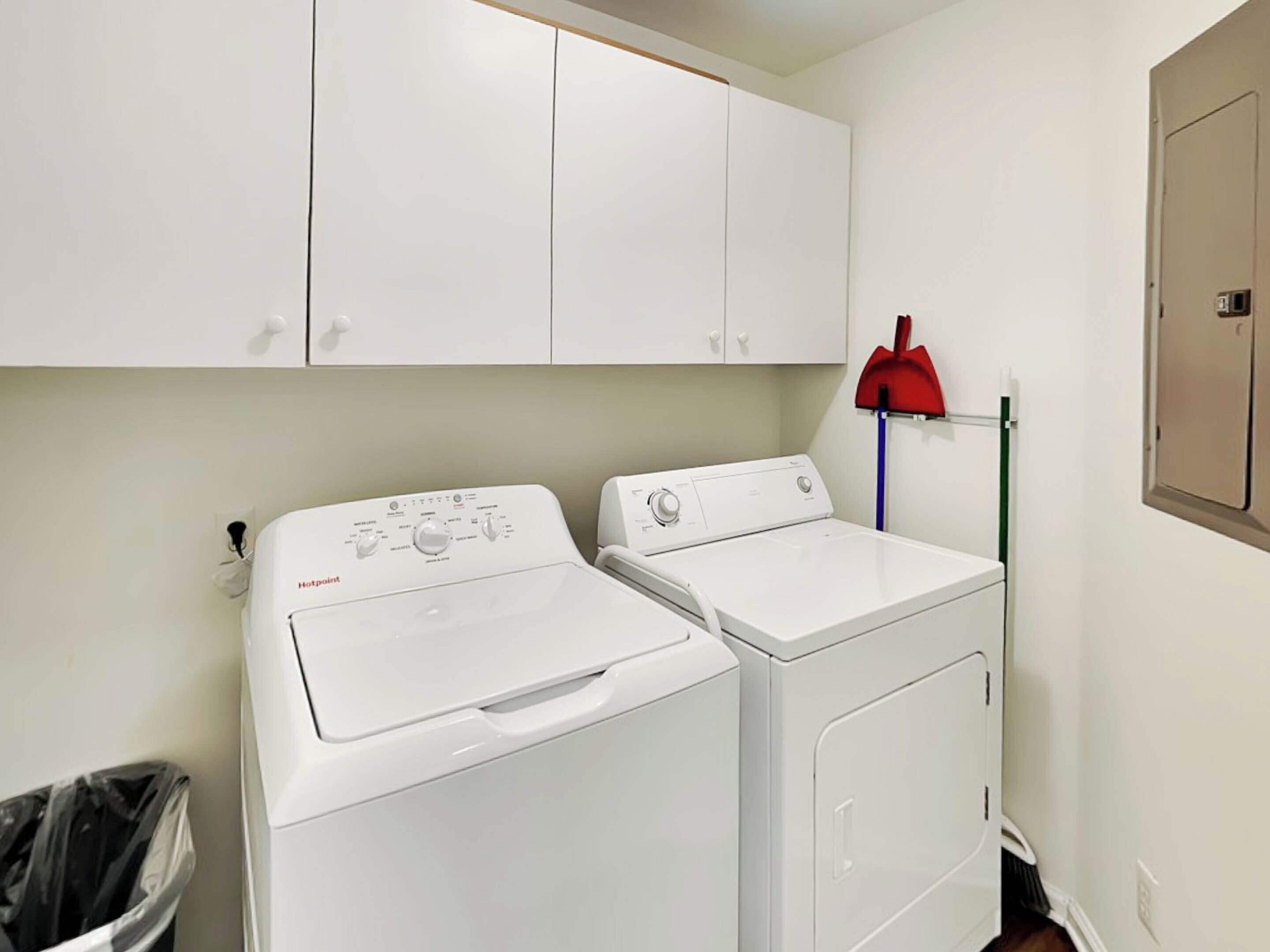 A laundry room with two white washing machines, a red broom, and a trash bin.