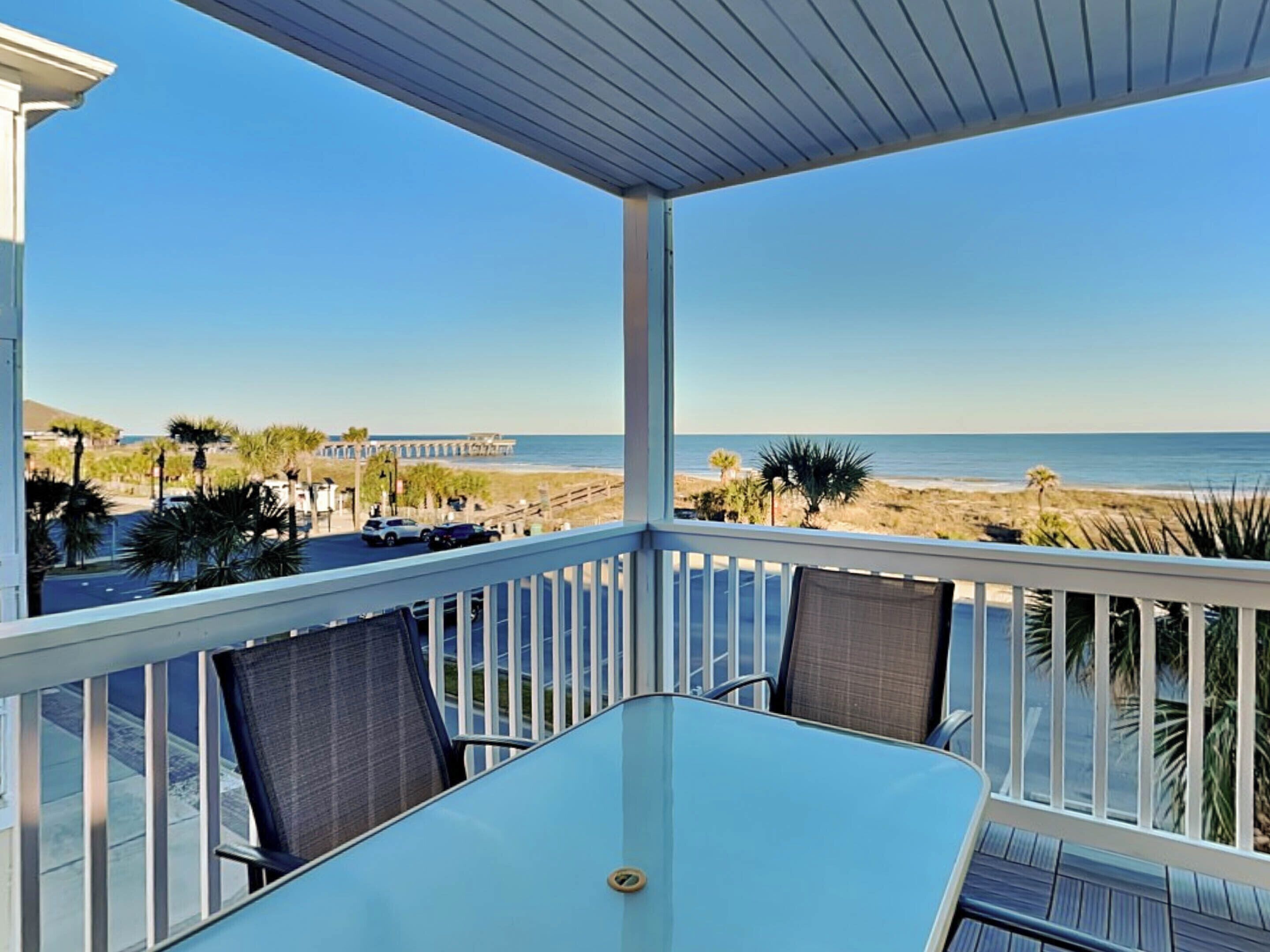 A balcony with a glass table and chairs, overlooking a beach and ocean.