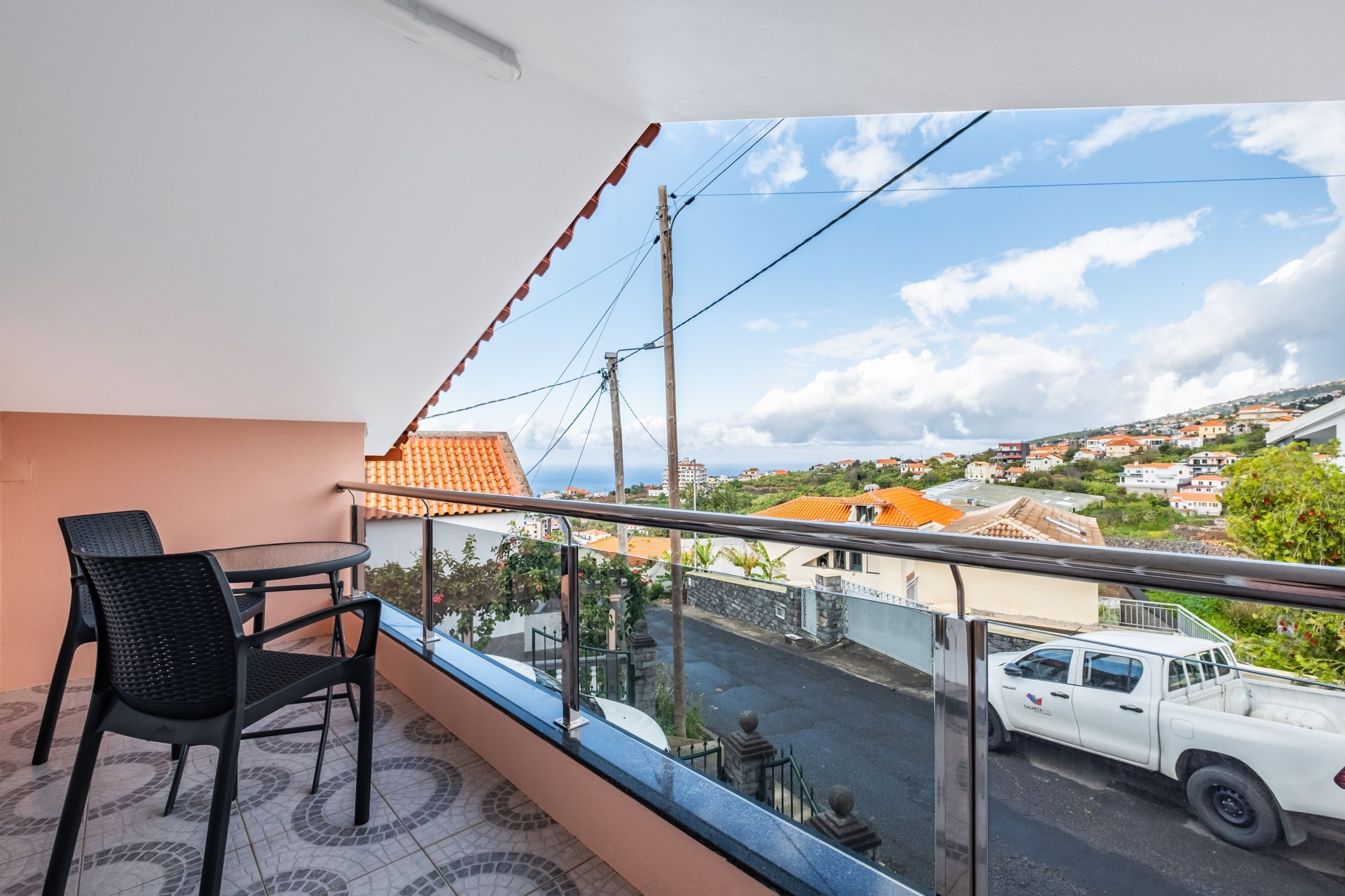 A balcony with a table and chairs, overlooking a street with houses and a white truck.