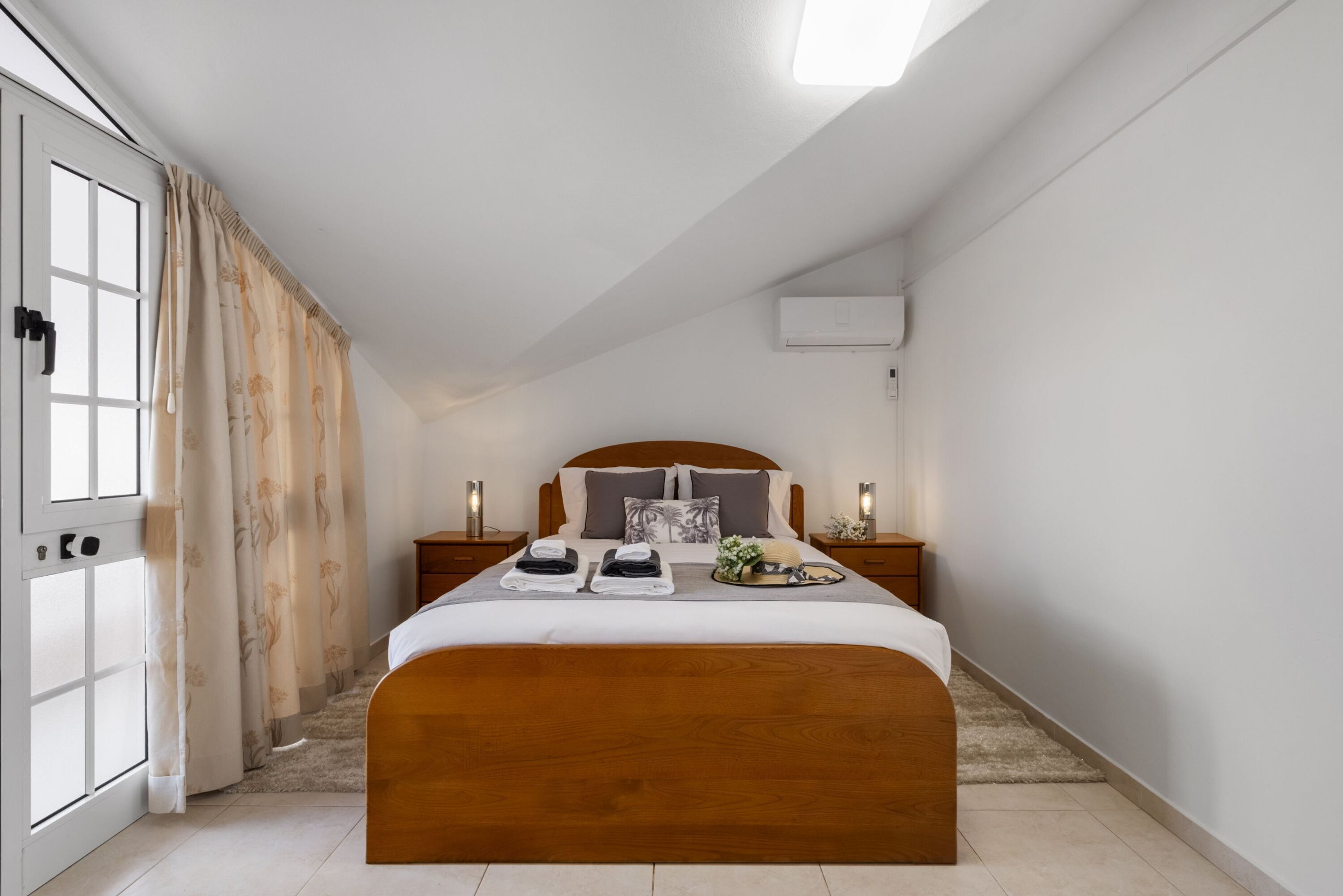 A bedroom with a wooden bed, a window with curtains, a bedside table with a lamp, and a wall-mounted air conditioner.