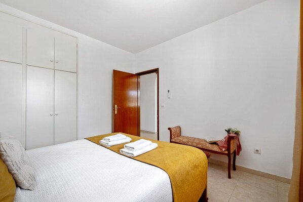 A bedroom with a bed, a wooden wardrobe, a bench, and a small table with a plant. - Golden Coast A by Homie (Calheta)