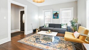 Un salon avec un canapé, une table basse et un fauteuil jaune. Il y a une grande fenêtre avec des stores, une porte et un luminaire suspendu.