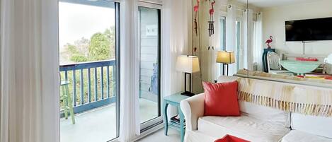 Una habitación con balcón, un sofá blanco, una mesita de tray en rojo y amarillo, y un televisor en la pared.