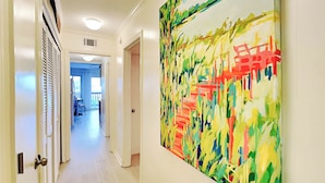 Un couloir avec une grande peinture abstraite au mur, une porte dotée d’une poignée et une vue sur une pièce avec une fenêtre.
