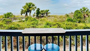 Une terrasse en bois avec deux chaises bleues, donnant sur une plage bordée de palmiers et surmontée d’une promenade en bois.