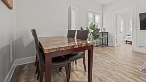 A dining area with a wooden table and chairs, a potted plant, and a view of a bedroom with a bed and a TV.