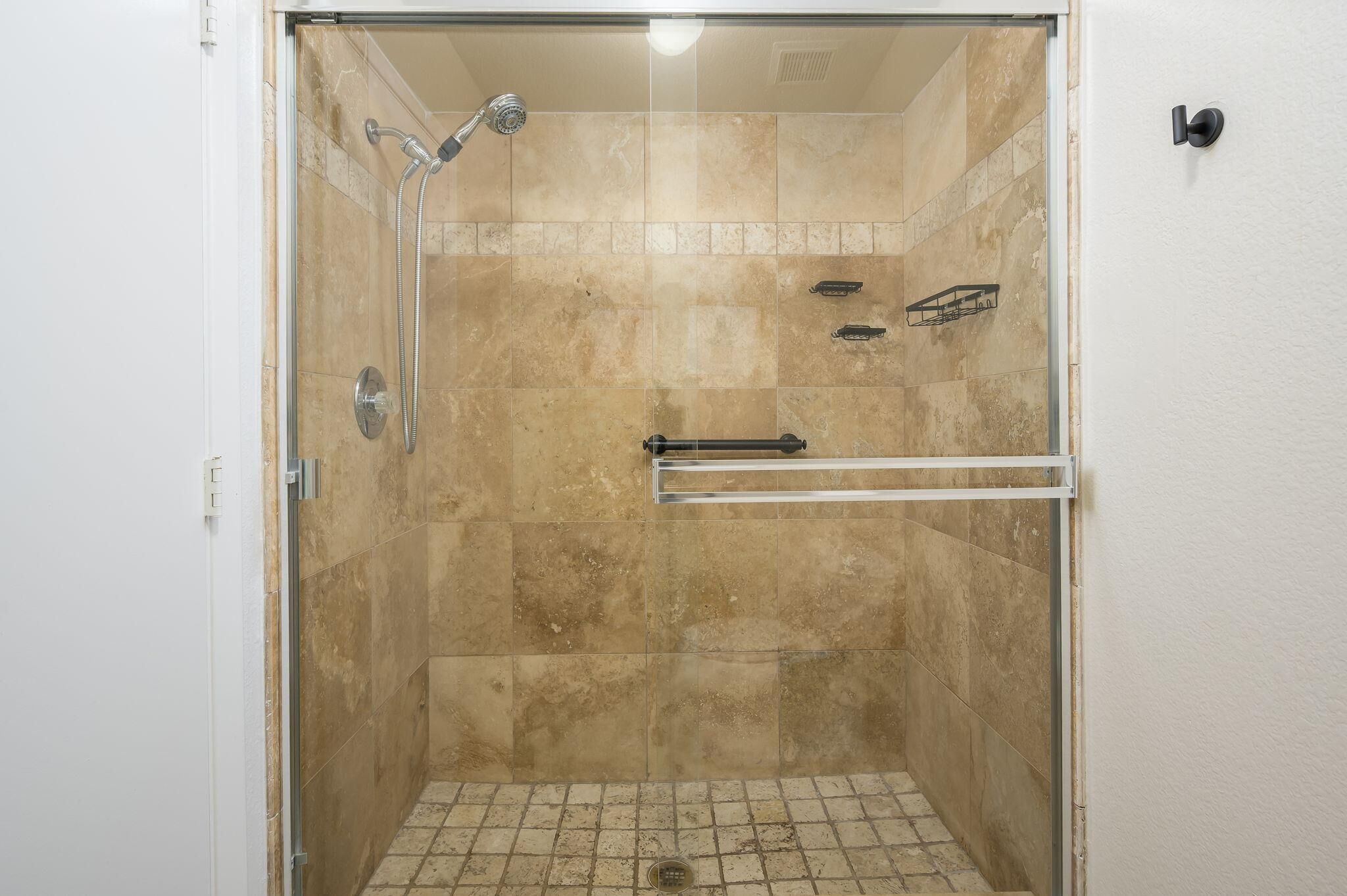 Une douche fermée par des parois de verre, avec des carreaux de couleur beige, une barre d’appui en métal et un drain visible.