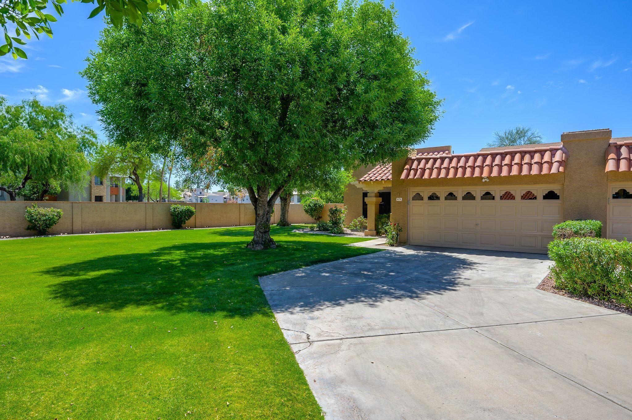 A residential property with a well-maintained lawn, a driveway leading to a garage, and a large tree providing shade.