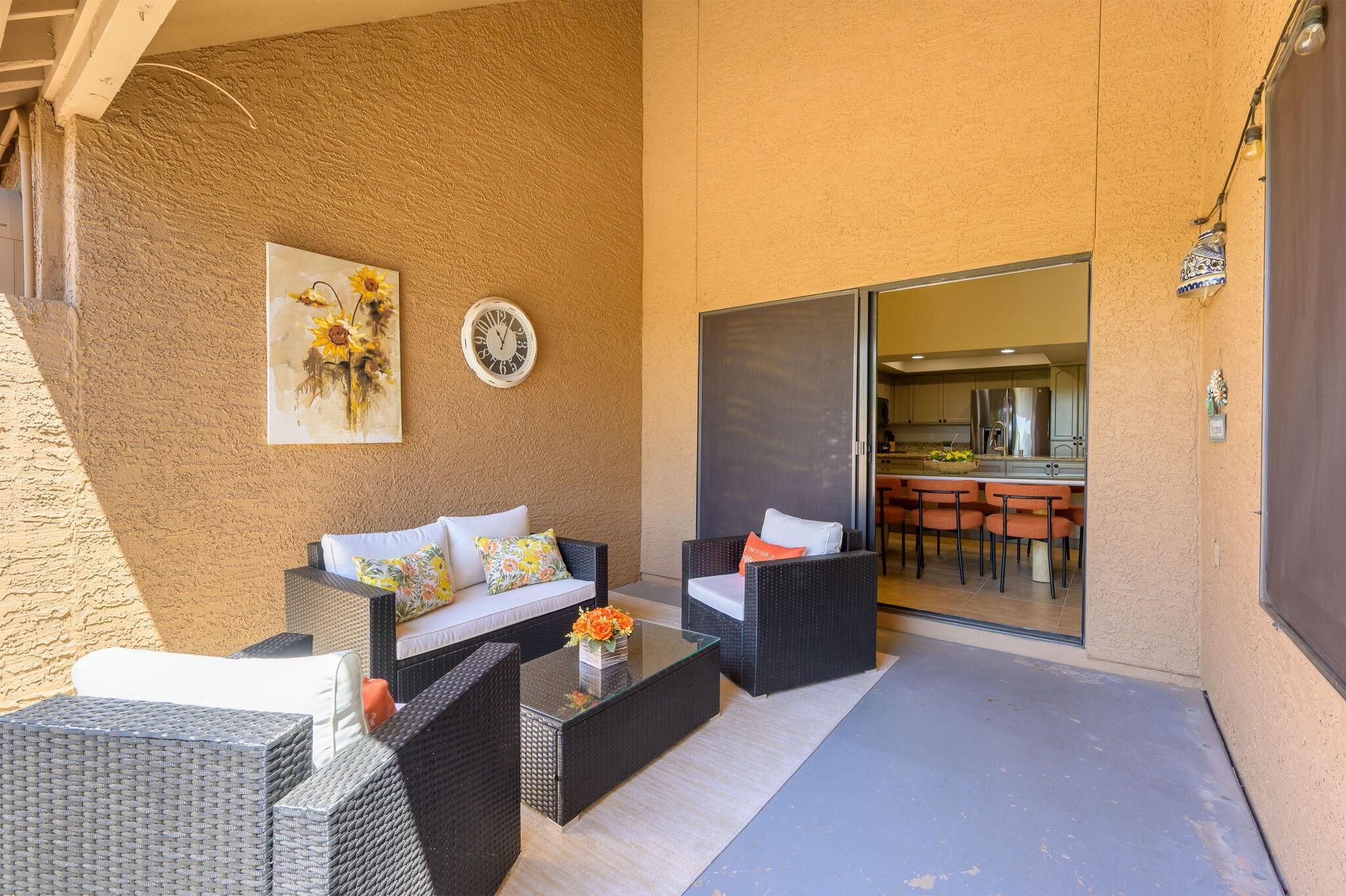 An outdoor patio area with wicker furniture, a glass coffee table, and a painting of sunflowers.