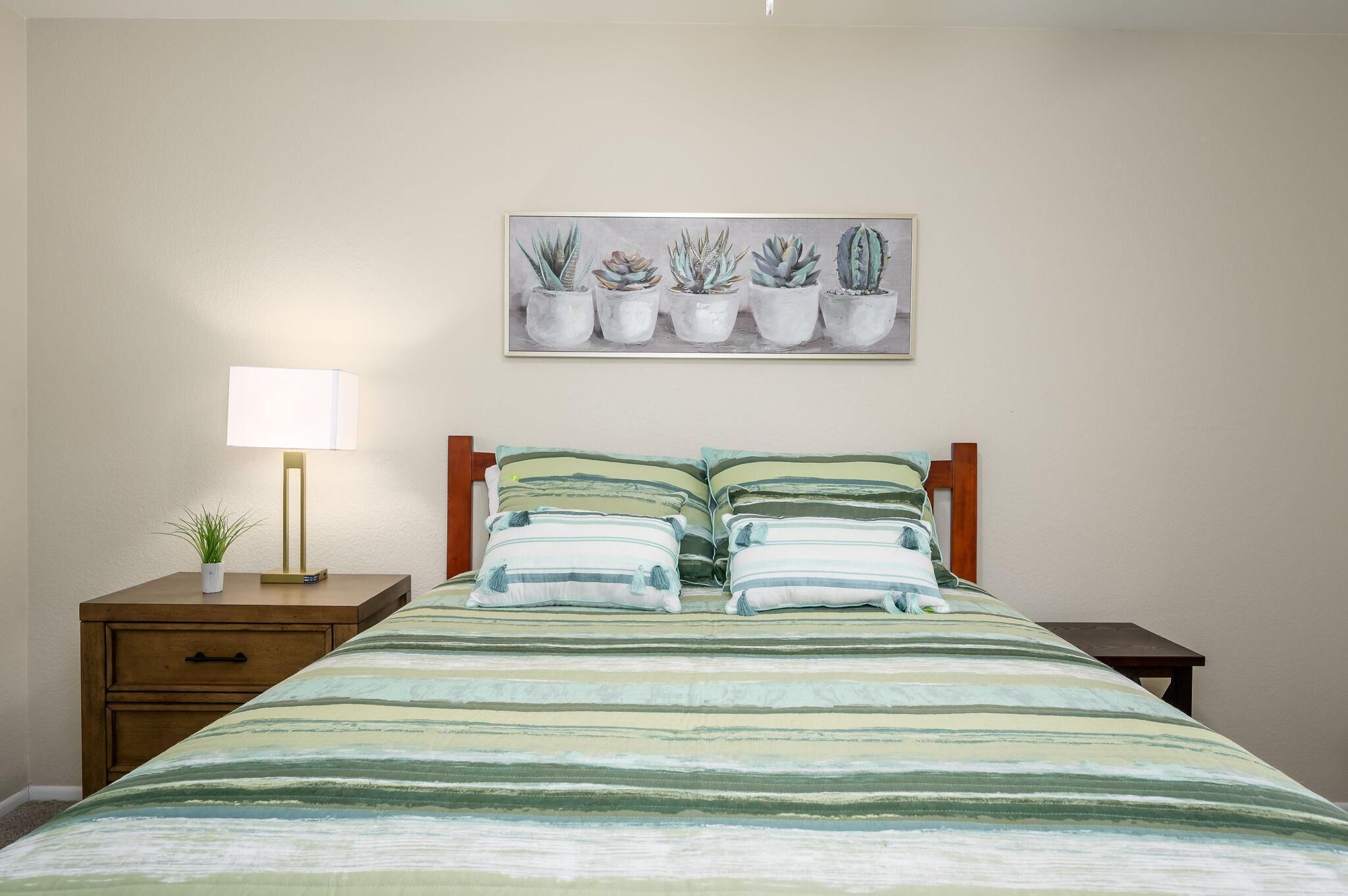 A bedroom with a bed, a nightstand with a lamp, a drawer, and a wall-mounted painting of potted plants.