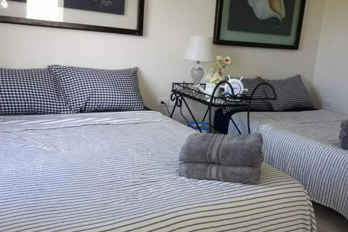 Habitación de hotel con dos camas, cada una con ropa de cama a rayas y almohadas cuadriculadas. Hay una mesita con una lámpara y un adorno.