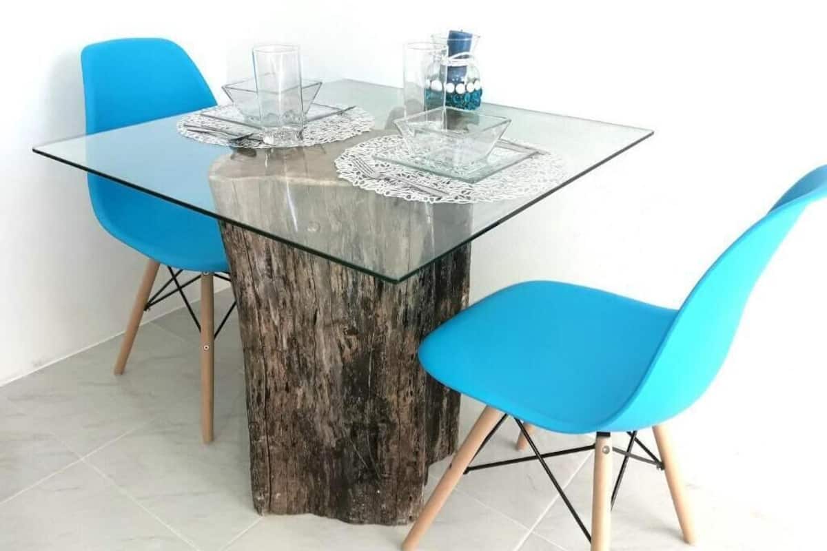 Una mesa de comedor con superficie de vidrio y base de madera, rodeada por dos sillas azules con patas de madera.