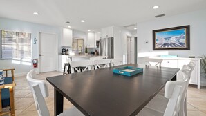 A modern kitchen with white cabinets, a stainless steel refrigerator, and a dining area with a dark wooden table and white chairs. - ⚓Mermaid Sunset! Coastal beach home w/pool 200 steps to beach!⚓ (Holmes Beach)