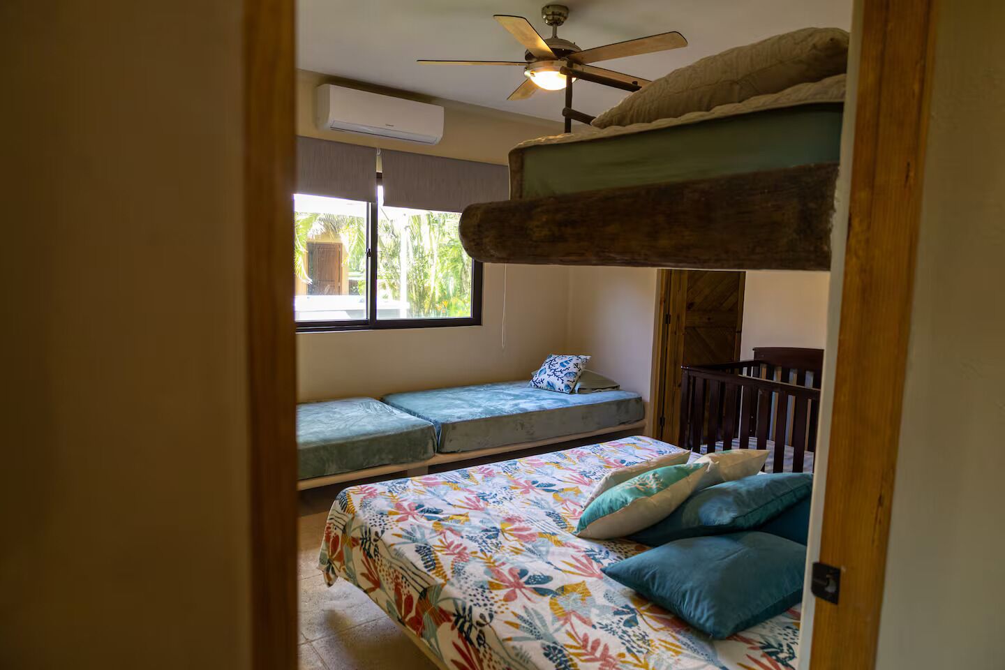 A bedroom with a bunk bed, a single bed, a ceiling fan, and a window with a view of palm trees.