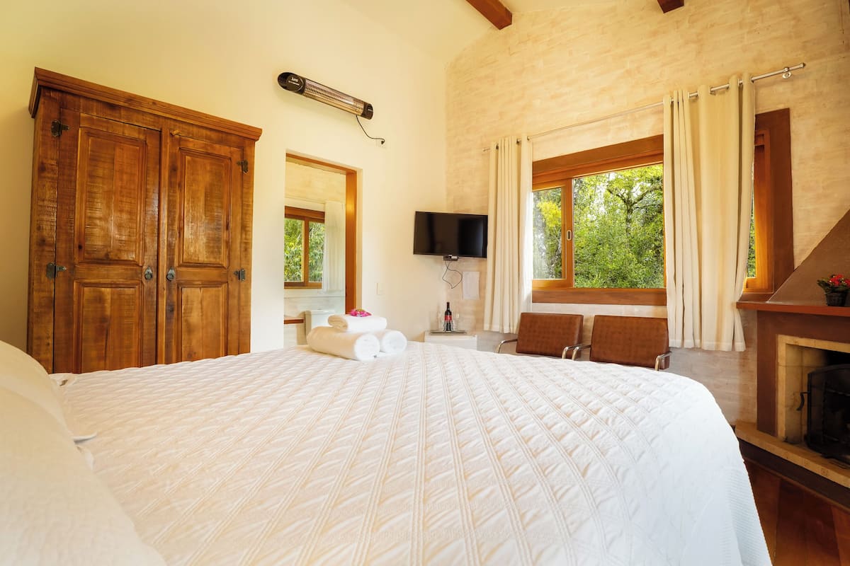 A bedroom with a large bed, wooden wardrobe, a flat-screen TV, a fireplace, and a window with curtains.