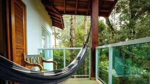 Una hamaca en un balcón con una silla de madera, una puerta de madera y vistas a un bosque frondoso.