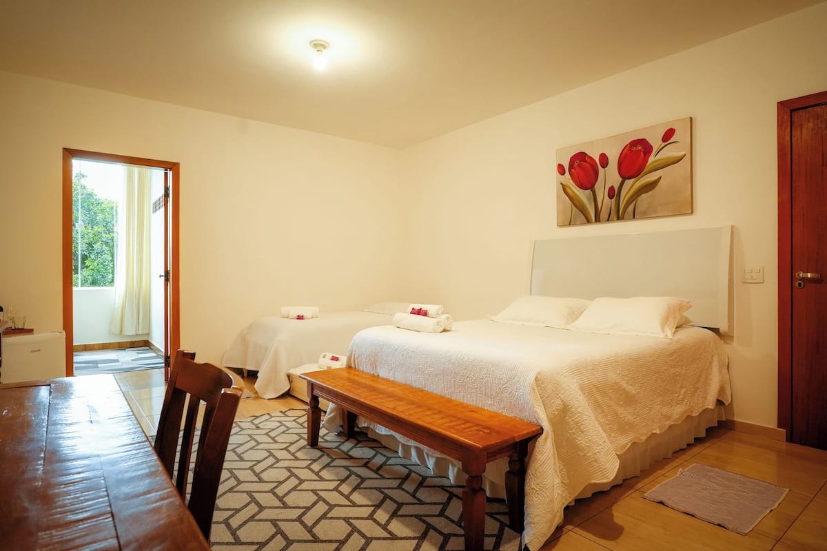 A bedroom with two beds, a wooden bench, a dining table, and a painting of red flowers on the wall.