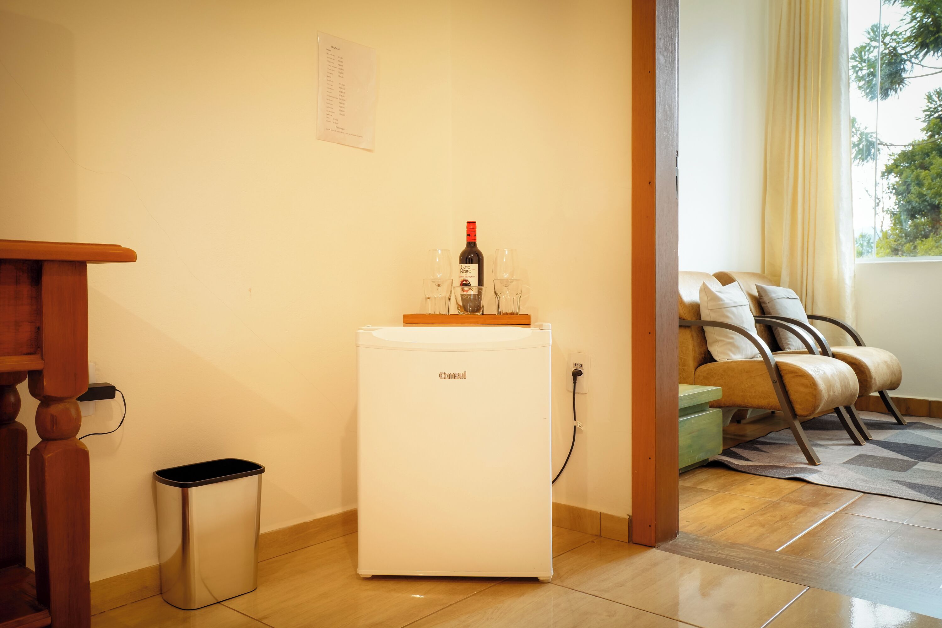 A compact room with a small white refrigerator, a trash bin, a wooden table, and a seating area with a sofa and cushions.