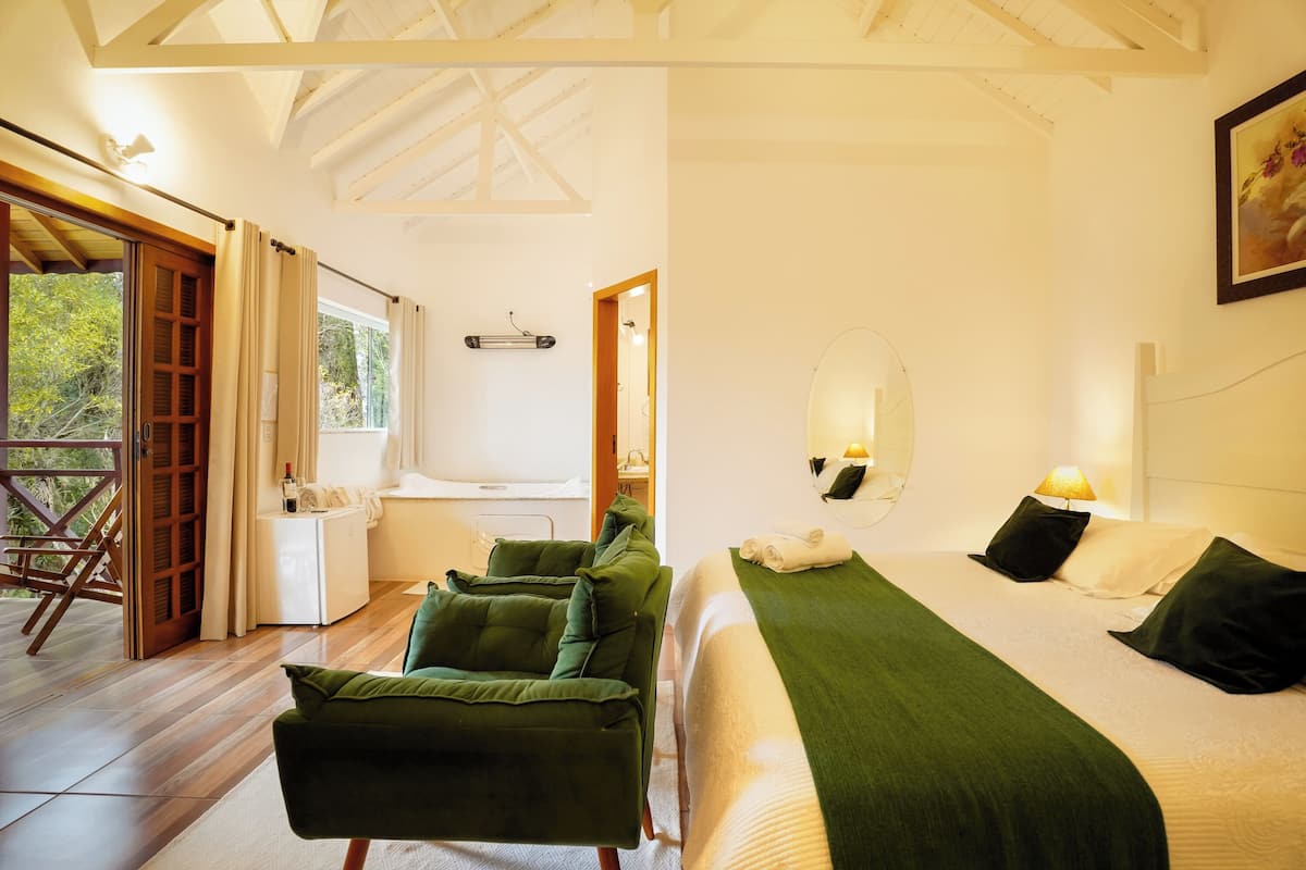 A bedroom with a bed, a green chair, a small table with a bottle, and a window.