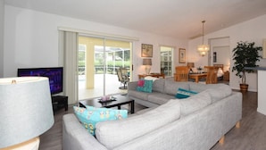 A modern living room with a sofa, coffee table, and TV. There is a dining area with a table and chairs, and a kitchen in the background.