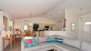 A living room with a grey sectional sofa, a coffee table, a dining area with a table and chairs, and a kitchen in the background.