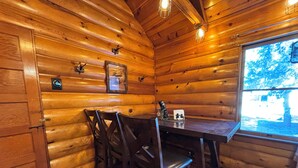 Interior de una cabaña de madera con una mesa de comedor y sillas, un ventanal con vista a árboles y una lámpara en el techo.
