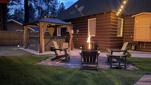 Una cabaña de troncos con un pozo de fuego, asientos exteriores y un patio cubierto.