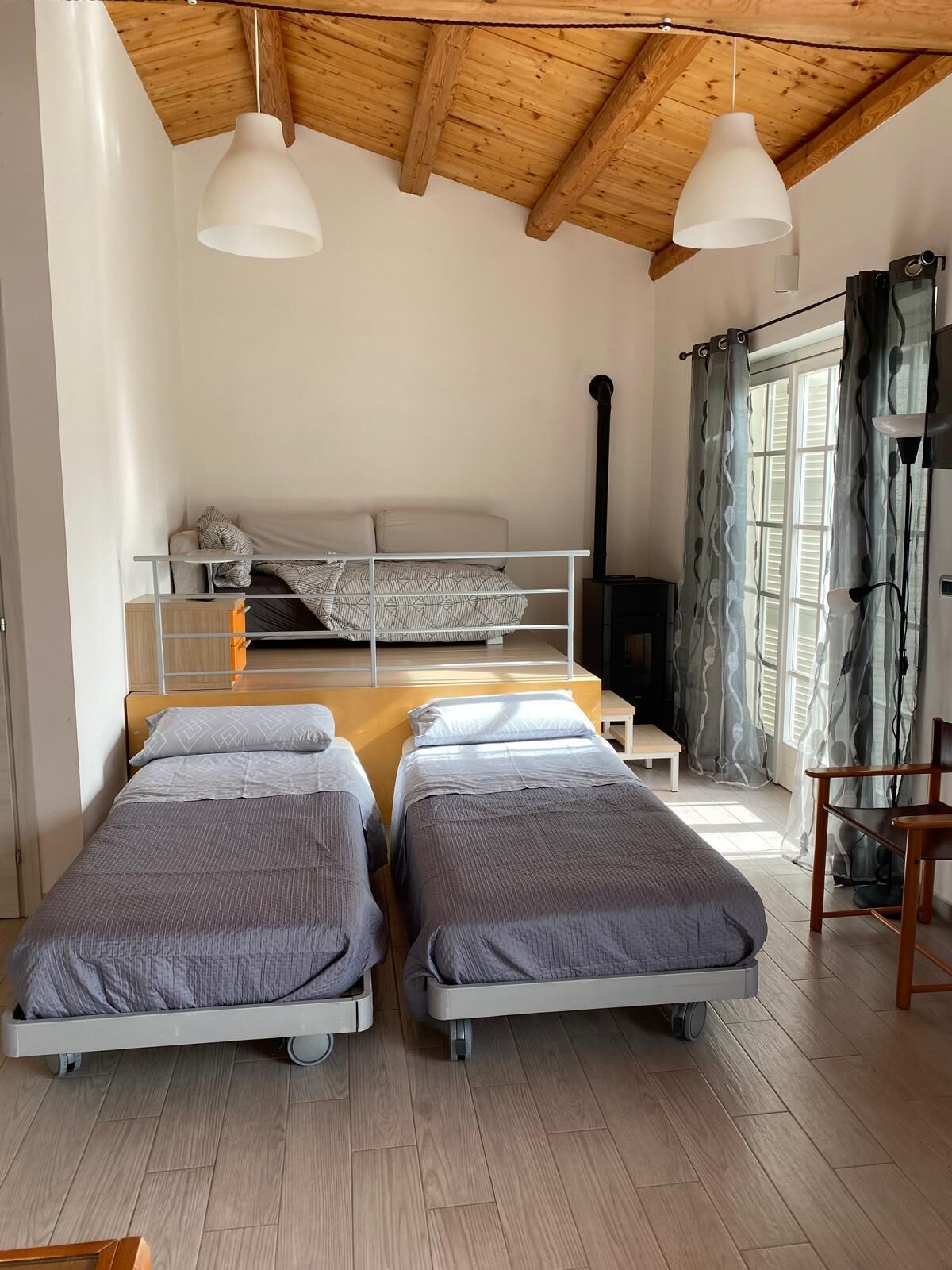 A room with two single beds, a bunk bed, a wooden ceiling, and a window with curtains.