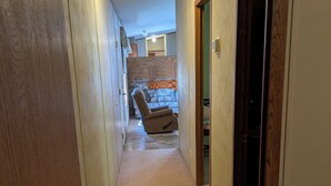 Un couloir étroit doté d’un sol recouvert de moquette, de murs en bois et d’une porte donnant sur une pièce où se trouvent une chaise et une petite table.