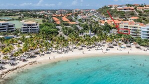 Vue aérienne d’un complexe hôtelier en bord de mer, avec une plage de sable, une eau turquoise et limpide, et plusieurs bâtiments en arrière-plan.