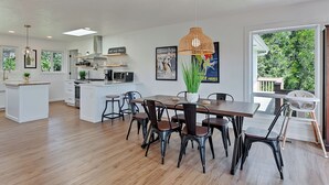 Una cocina moderna con armarios blancos, una isla central y una zona de comedor con mesas y sillas de madera.