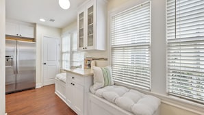 Una cocina con muebles blancos, un banco empotrado con cojines y una ventana con persianas.