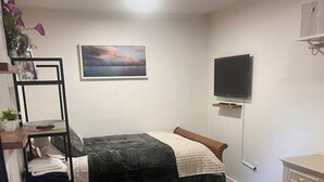 A bedroom with a bed, a desk, a chair, a TV, and a shelf with folded clothes.