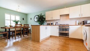 A modern kitchen with wooden flooring, white cabinets, a dining area with chairs, and a large window.
