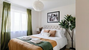 A neatly made bed with a green and white blanket, a framed photograph on the wall, and a potted plant beside the bed.