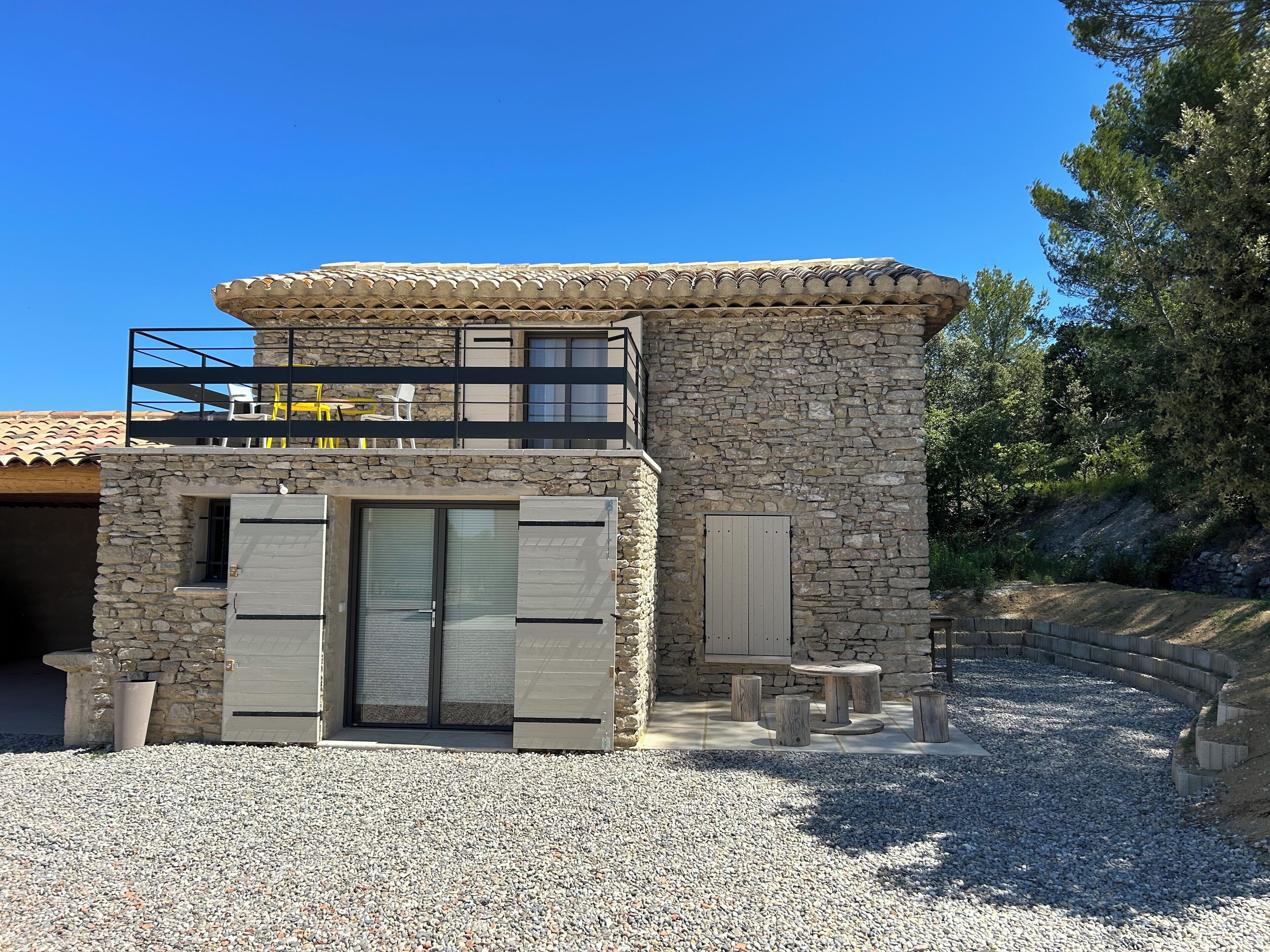 A two-story stone building with a balcony, a door with shutters, and a gravel driveway.