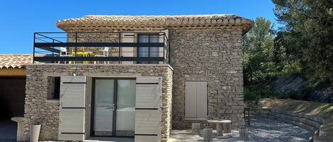 A two-story stone building with a balcony, a door with shutters, and a gravel driveway.