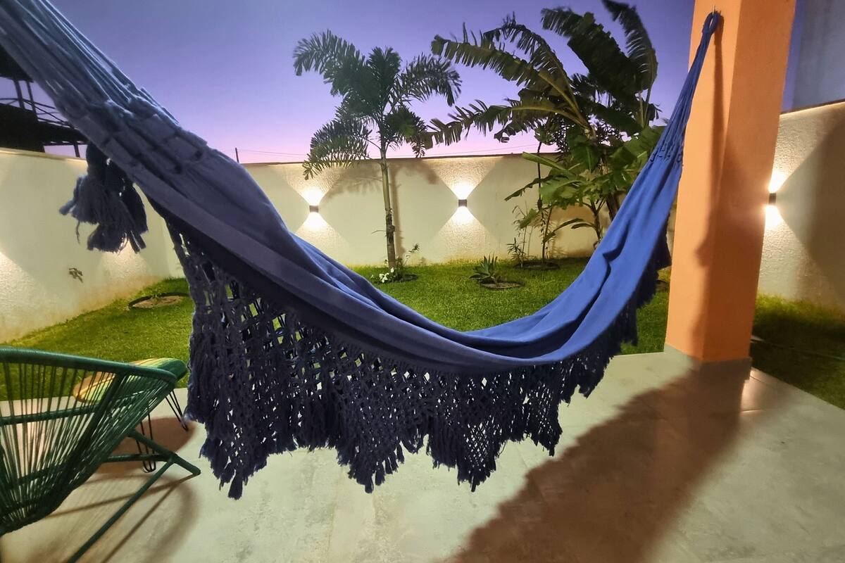 Un hamac suspendu entre deux poteaux, une chaise verte et un petit jardin avec des palmiers et un mur décoré de guirlandes lumineuses.