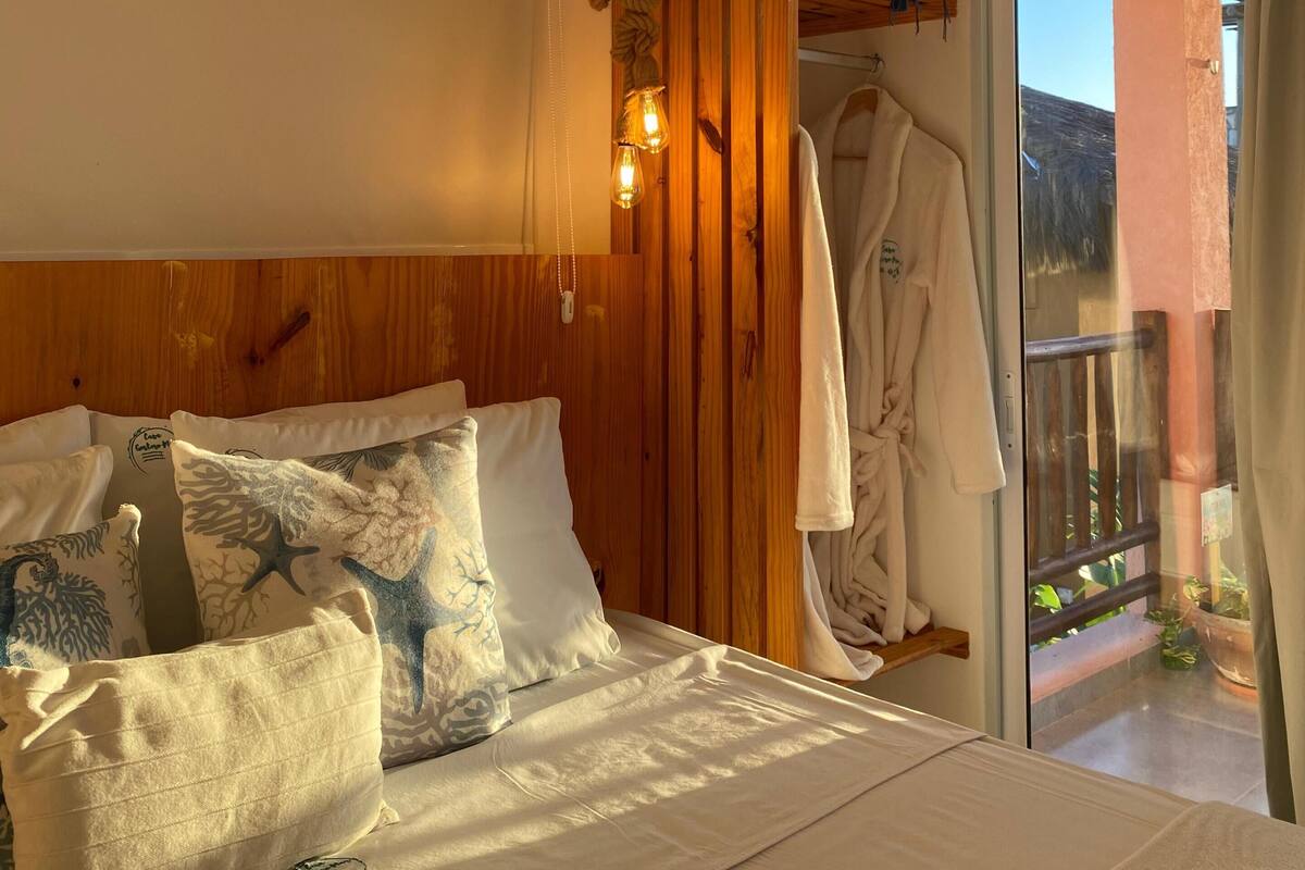 Une chambre avec une tête de lit en bois, un lit agrémenté d’oreillers à motifs, un peignoir suspendu à un porte-manteau et une vue sur l’extérieur par une porte vitrée.