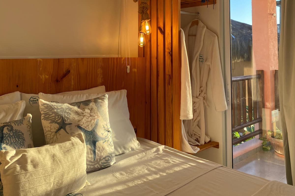 A bedroom with a wooden headboard, a bed with patterned pillows, a robe hanging on a rack, and a view of the outdoors through a glass door.