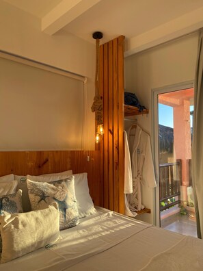 Une chambre avec une tête de lit en bois, un lit agrémenté d’oreillers à motifs, un peignoir suspendu à un porte-manteau et une vue sur l’extérieur par une porte vitrée.