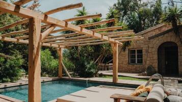 A wooden pergola structure over a swimming pool with lounge chairs and a stone building in the background.