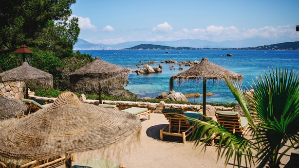 A beachside lounge with thatched umbrellas and wooden lounge chairs, overlooking a clear blue sea and rocky coastline.