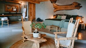 A rustic room with a large wooden headboard bed, a wooden chair, a small table with a potted plant, and a wooden sculpture on the wall.