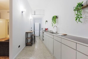 A narrow kitchen with white cabinets, a countertop, and a sink. There are two stools and a small table.