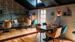 Un espace repas doté d’un plancher en bois, d’un plafond haut et d’une grande fenêtre. On y trouve des chaises et une table sur laquelle est posé un vase rempli de fleurs.